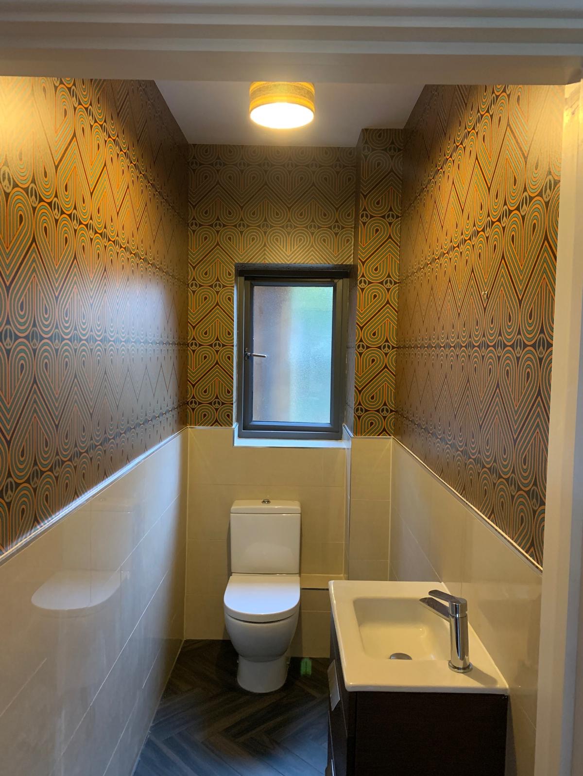 Wąska łazienka po remoncie z toaletą, umywalką na szafce i wzorzystą tapetą w stylu retro na górnej części ścian.