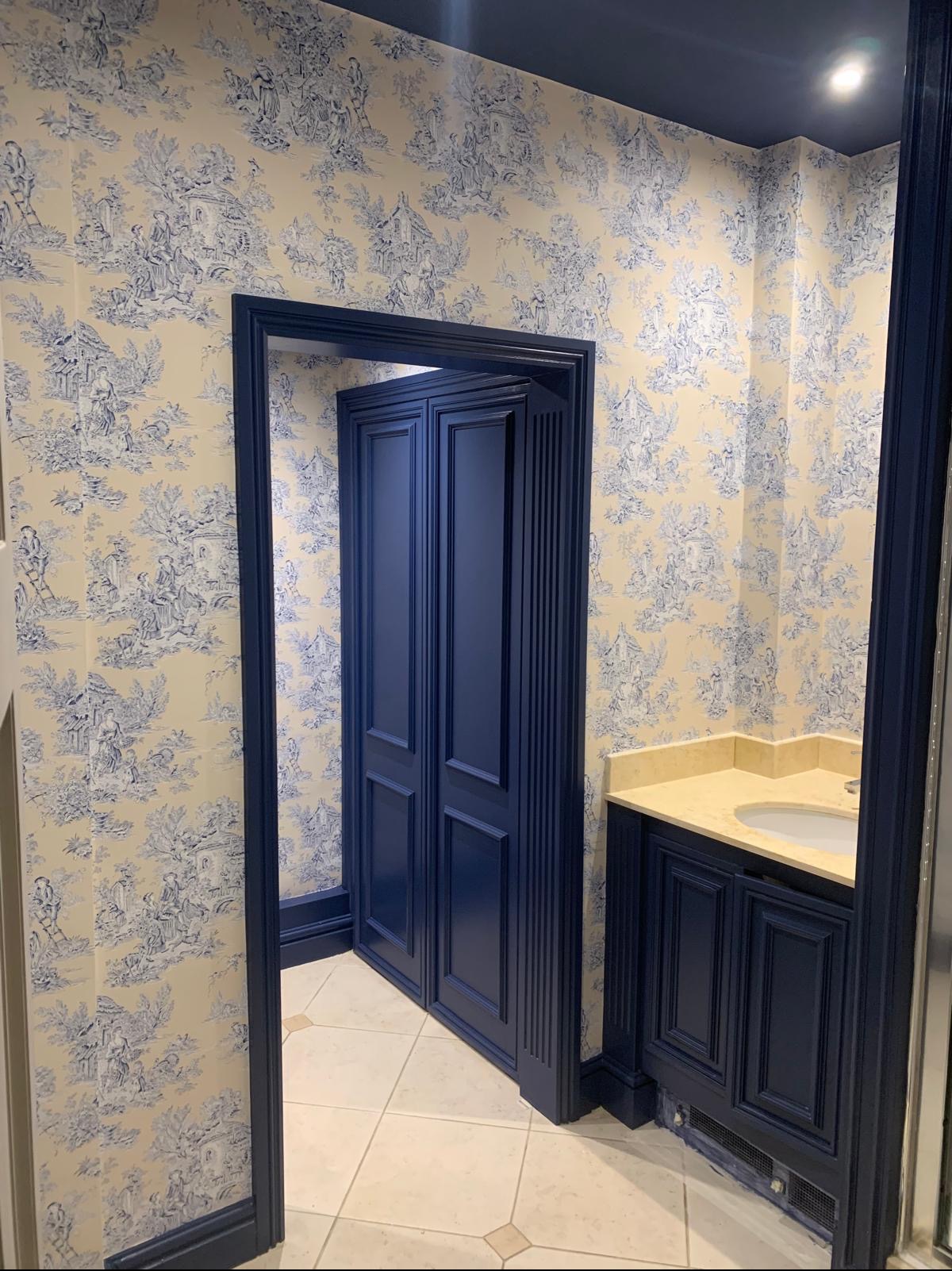 Elegancka łazienka z granatowymi drzwiami i szafką umywalkową, ściany pokryte tapetą w stylu toile de jouy, beżowa umywalka i płytki podłogowe.
