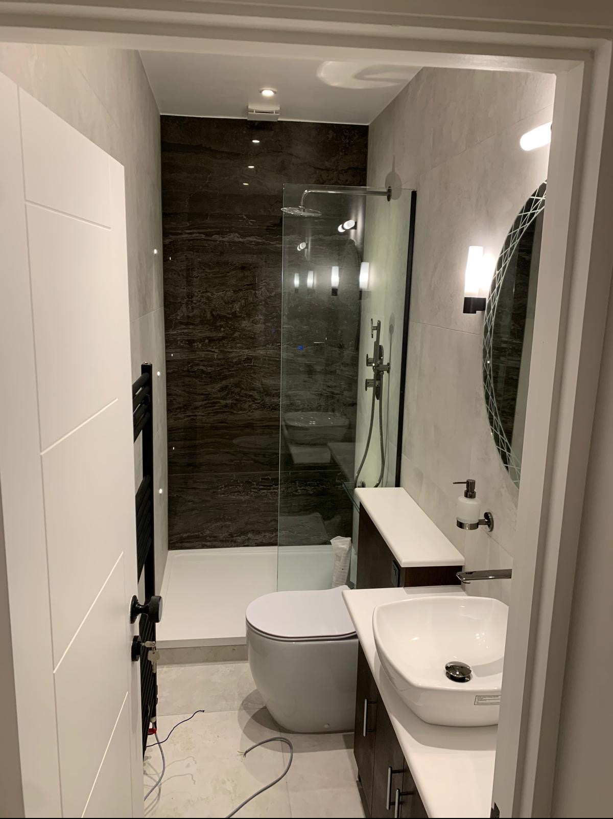 Nowoczesna łazienka z kabiną prysznicową typu walk-in, umywalką nablatową i dużym owalnym lustrem, wykończona szarymi płytkami i akcentami z ciemnego marmuru.