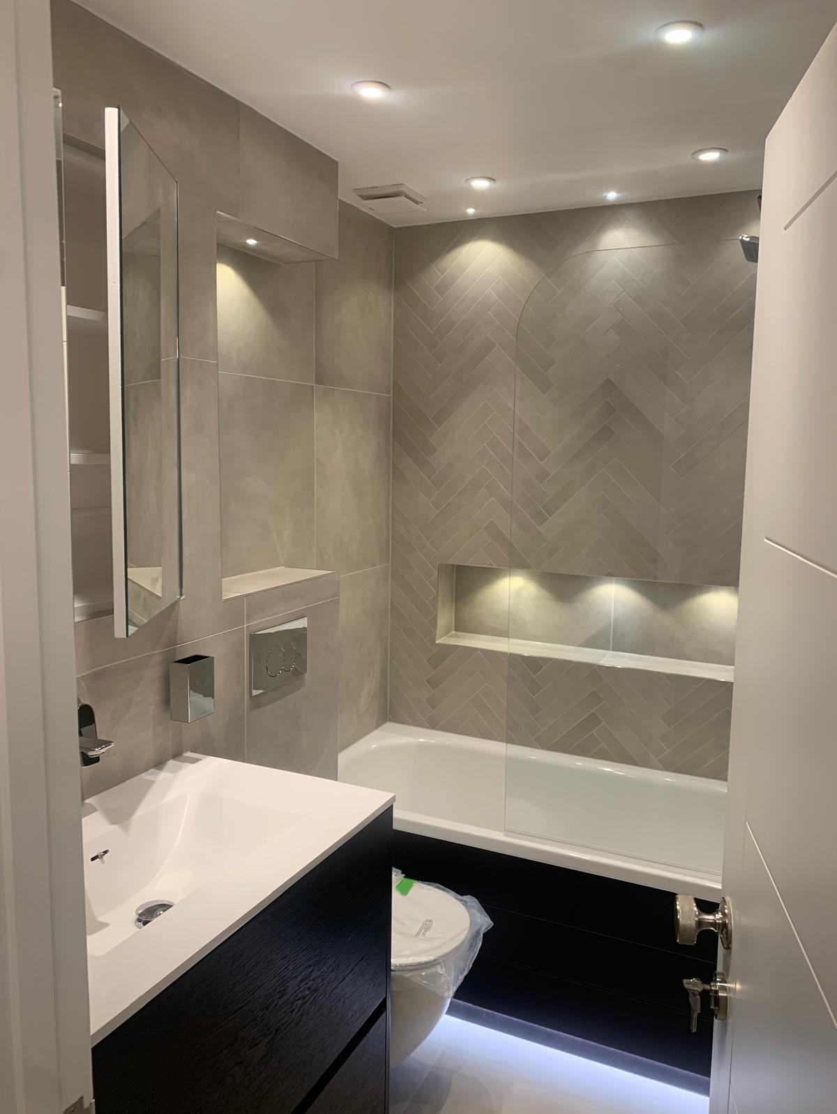 Nowoczesna łazienka po remoncie z beżowymi płytkami ułożonymi w jodełkę, podświetlaną wnęką nad wanną, szafką z umywalką w kolorze czarnym, lustrem z półkami i toaletą w folii ochronnej.
