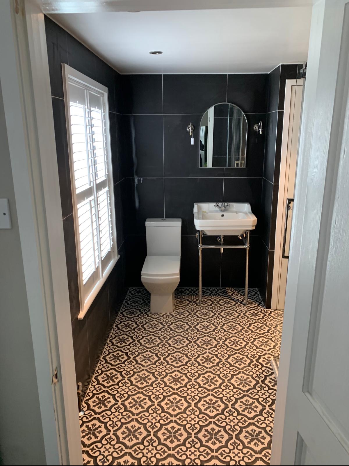 Nowoczesna łazienka z czarnymi płytkami ściennymi, wzorzystą czarno-białą podłogą, białą toaletą i umywalką na chromowanym stelażu, oświetlona przez okno z żaluzjami.