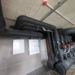 Czarno izolowane rury instalacji grzewczej z czerwonymi miernikami, białe skrzynki sterownicze na szarej ścianie, sufit z elementami instalacji podwieszanej.