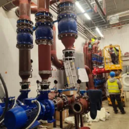 Rozbudowana instalacja hydrauliczna z rurami w kolorach brązowym i czerwonym, łączona niebieskimi elementami, widoczni pracownicy na podnośniku i na ziemi, narzędzia na stole.