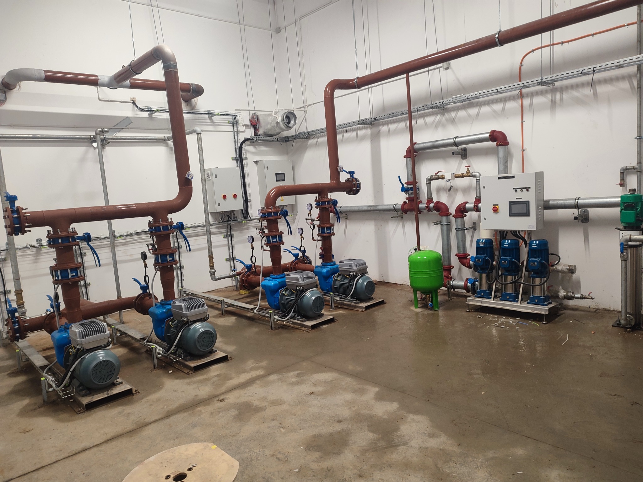 Kompleksowa instalacja pompowni z rzędami niebieskich pomp i brązowymi rurami, widoczna szafa sterownicza i zielony zbiornik wyrównawczy.