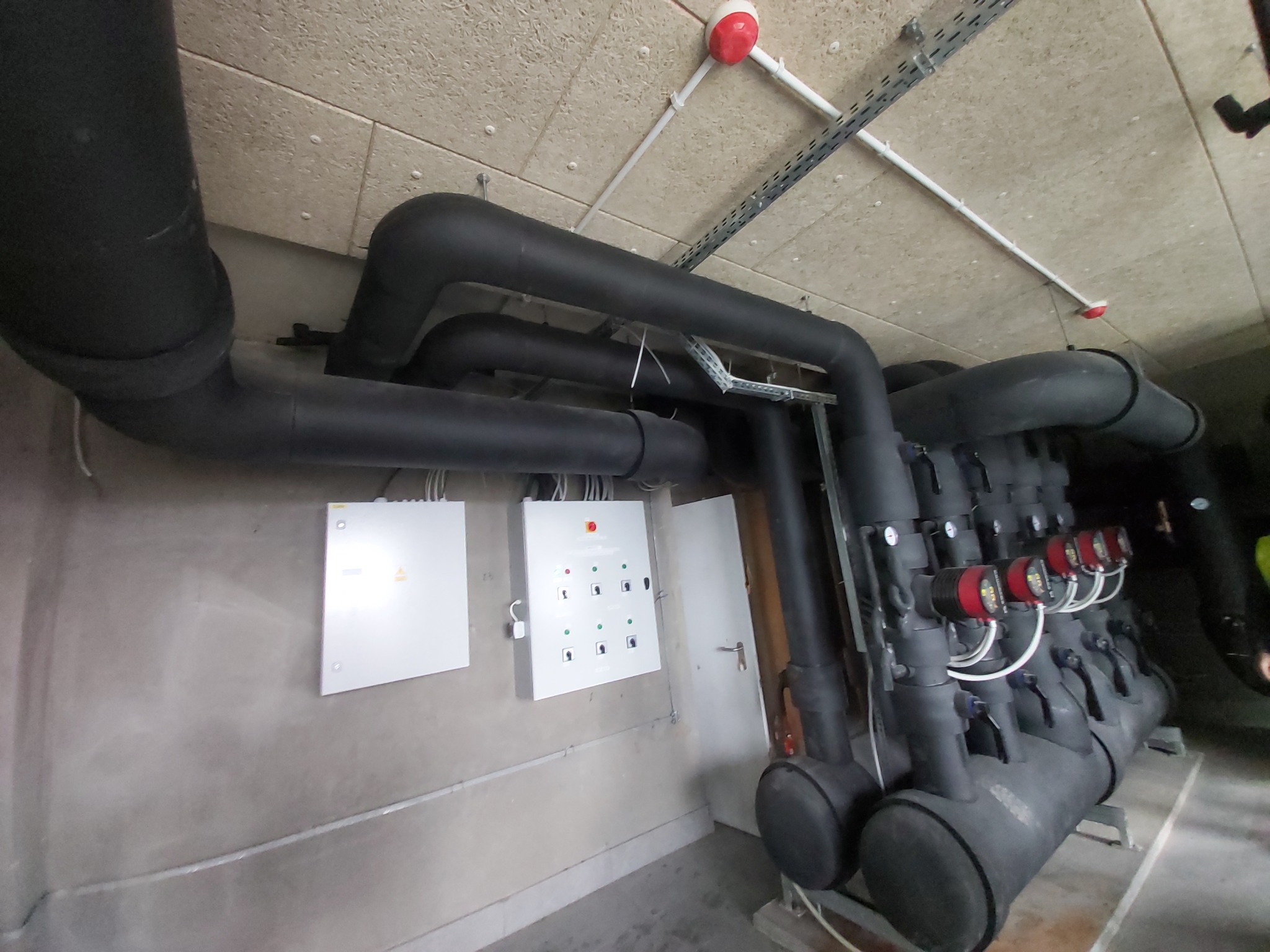 Czarno izolowane rury instalacji grzewczej z czerwonymi miernikami, białe skrzynki sterownicze na szarej ścianie, sufit z elementami instalacji podwieszanej.