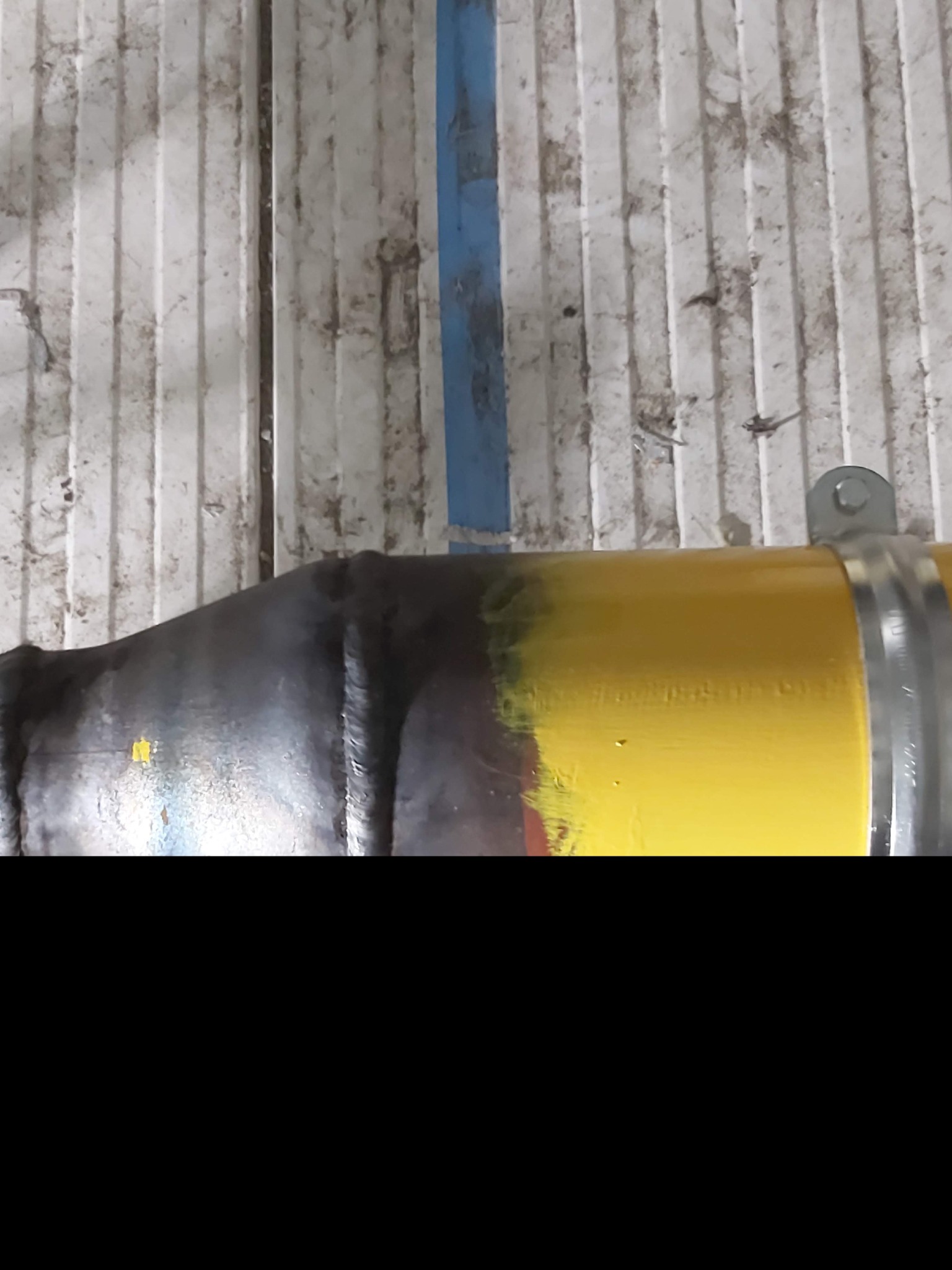 Żółta rura gazowa z metalową obejmą przymocowana do brudnej, białej ściany z niebieskim paskiem.