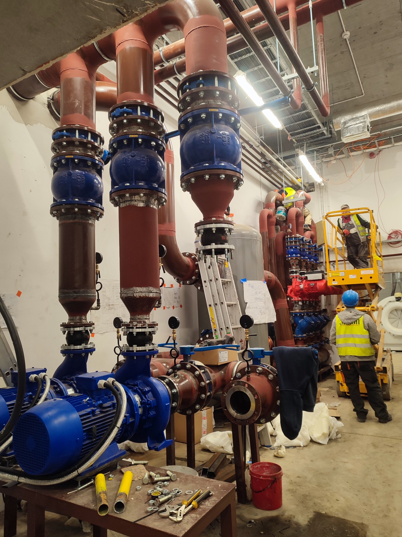 Rozbudowana instalacja hydrauliczna z rurami w kolorach brązowym i czerwonym, łączona niebieskimi elementami, widoczni pracownicy na podnośniku i na ziemi, narzędzia na stole.