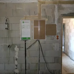 Surowa ściana z betonu komórkowego w trakcie prac elektrycznych: widoczne kable prowadzone po ścianie, puszki elektryczne, tablica rozdzielcza Elektro-Plast MSF RP 4/56 i prowizoryczne oświetlenie...
