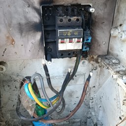 Elektryczne Innowacje - Uszkodzona skrzynka elektryczna w Chwaszczynie z przepalonymi bezpiecznikami i okopconymi ściankami, widoczne kable i ślady po zwarciu.