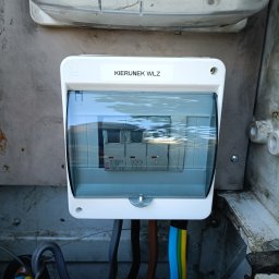 Elektryczne Innowacje - Skrzynka elektryczna z bezpiecznikami za przezroczystą osłoną, z naklejką 'KIERUNEK WLZ' i widocznymi przewodami.