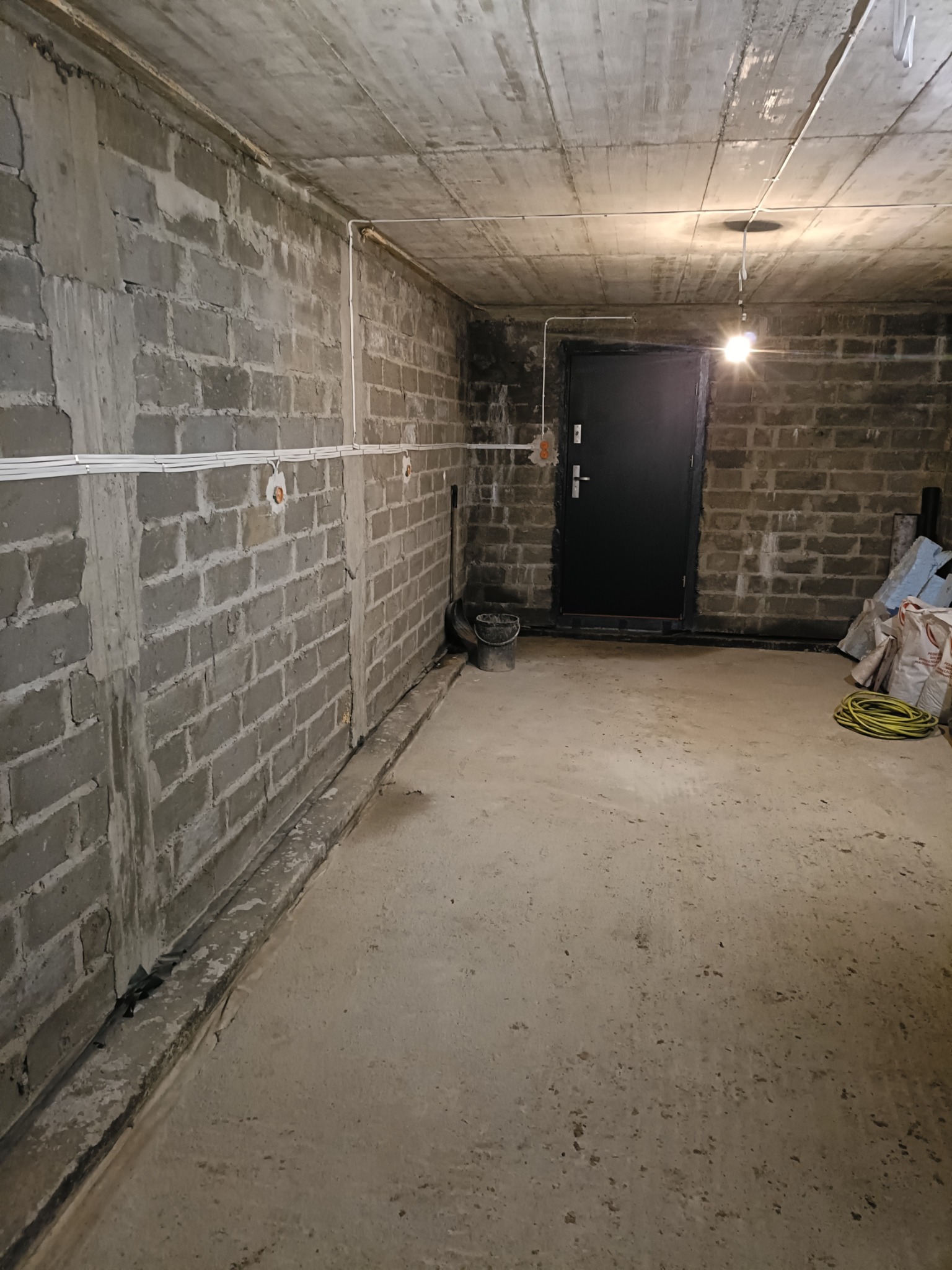 Surowy stan piwnicy z widocznymi przewodami elektrycznymi przymocowanymi do ściany z betonowych pustaków, pojedyncza żarówka oświetla przestrzeń, czarne drzwi w tle.