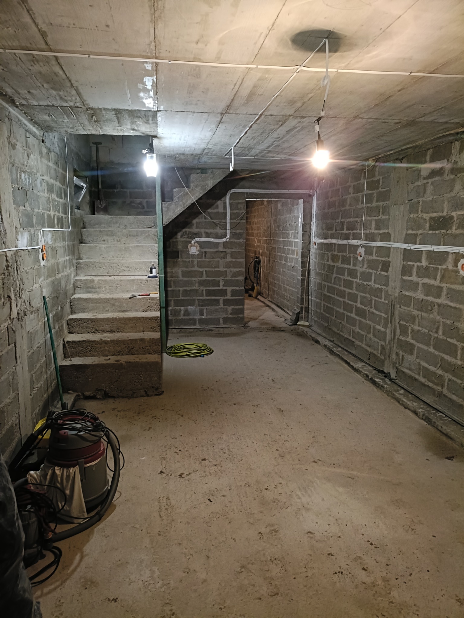 Surowe wnętrze piwnicy z betonowymi ścianami, schodami i widocznymi przewodami elektrycznymi pod sufitem, oświetlone wiszącymi żarówkami. Na podłodze leży zwinięty kabel i stoi odkurzacz przemysłowy.