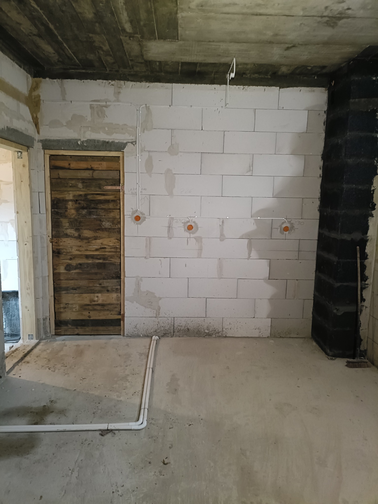 Surowy stan wnętrza z widocznymi przewodami elektrycznymi poprowadzonymi po ścianie z bloków, puszkami instalacyjnymi i prowizorycznymi drzwiami z desek.