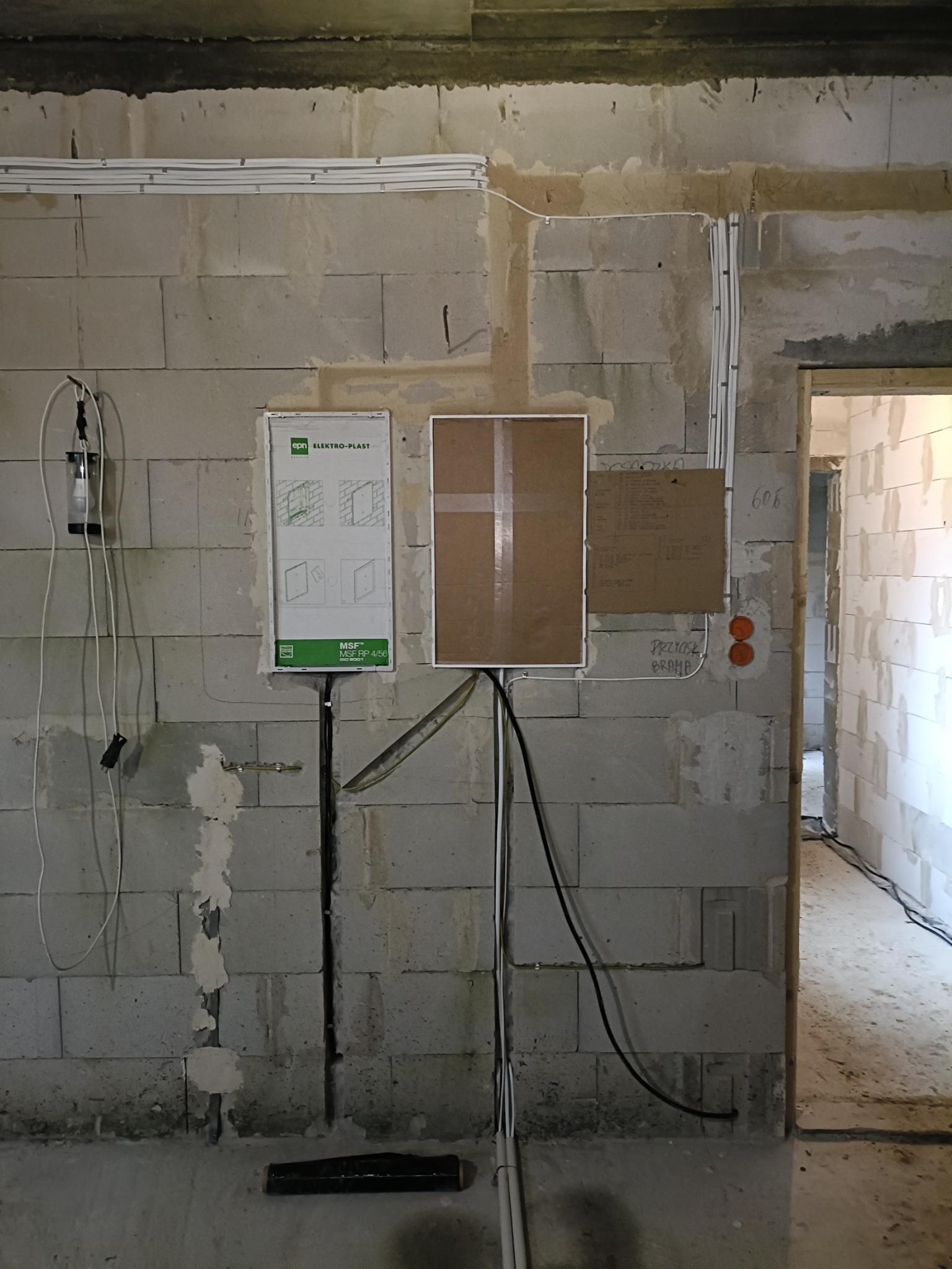 Surowa ściana z betonu komórkowego w trakcie prac elektrycznych: widoczne kable prowadzone po ścianie, puszki elektryczne, tablica rozdzielcza Elektro-Plast MSF RP 4/56 i prowizoryczne oświetlenie...