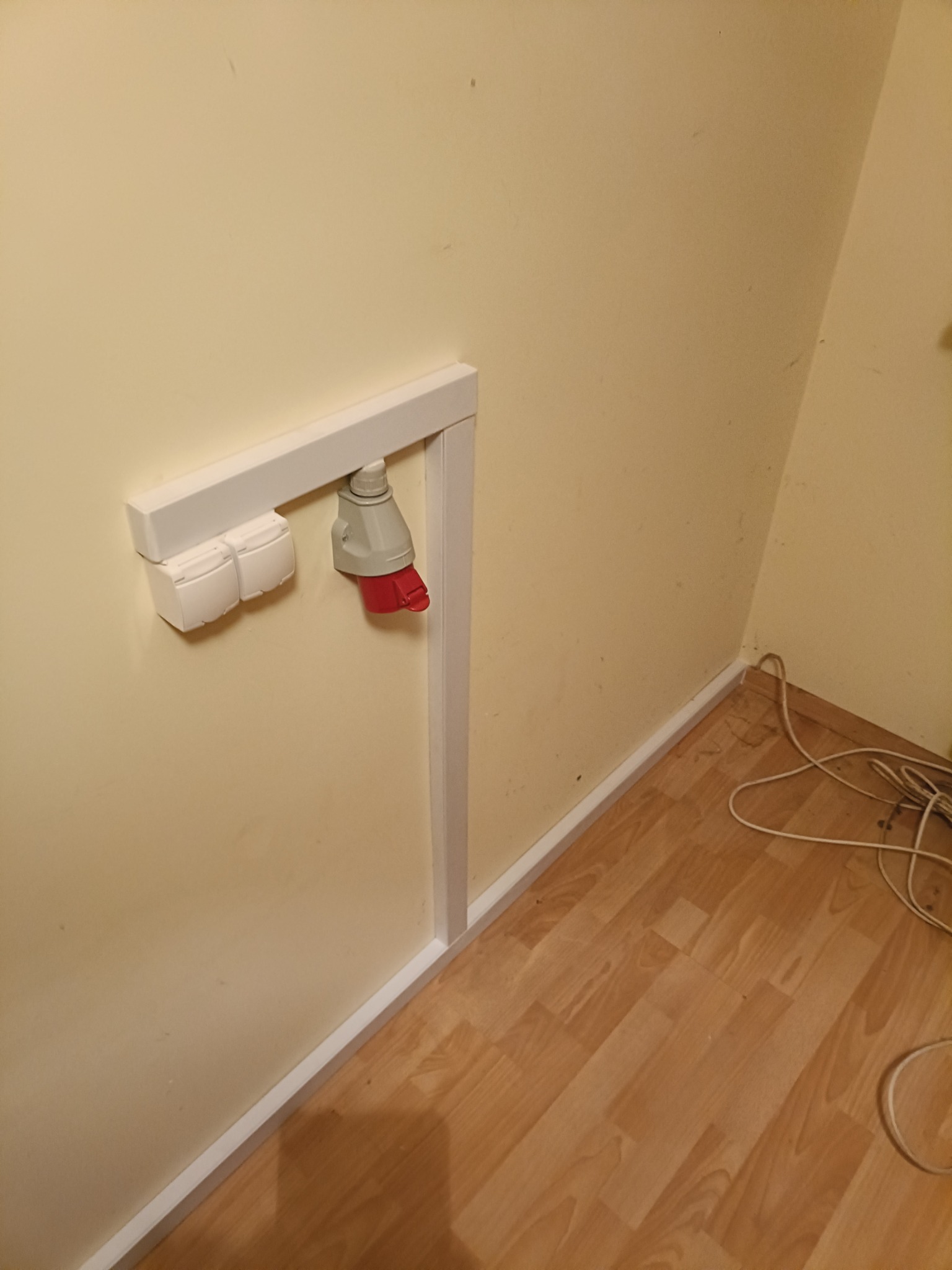 Zamontowane na ścianie gniazdo siłowe z wtyczką przemysłową, obudowane białą ramką ochronną.