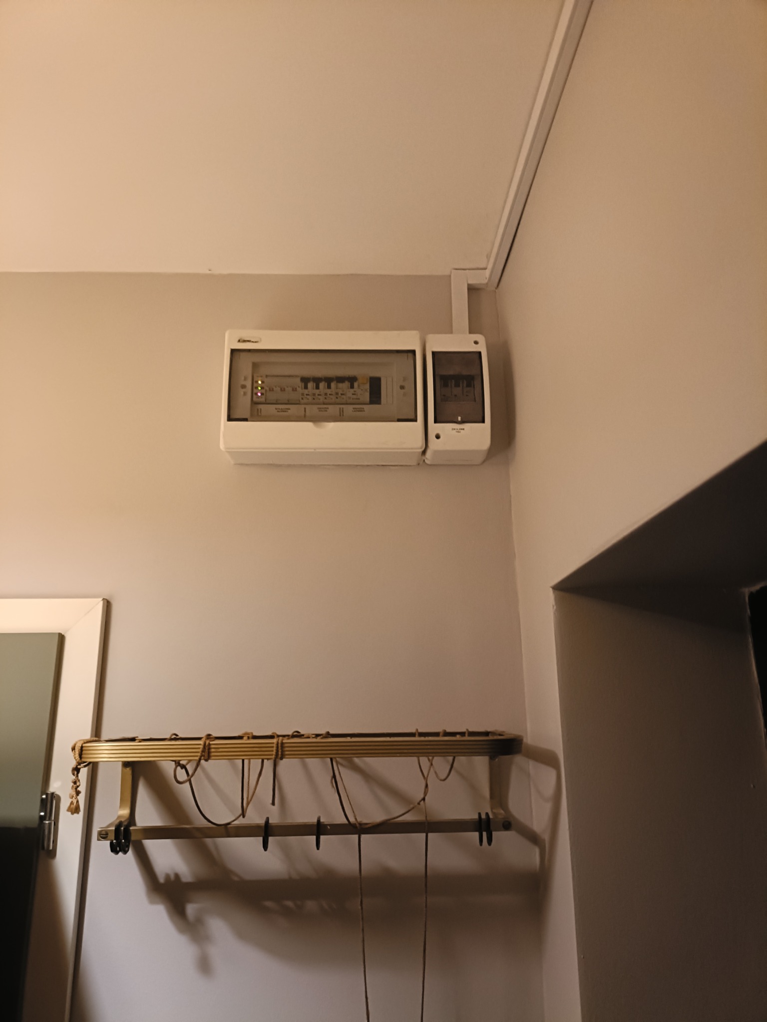 Dwa białe panele elektryczne zamontowane na szarej ścianie nad wieszakiem na ubrania, widoczne przewody i zabezpieczenia wewnątrz paneli.