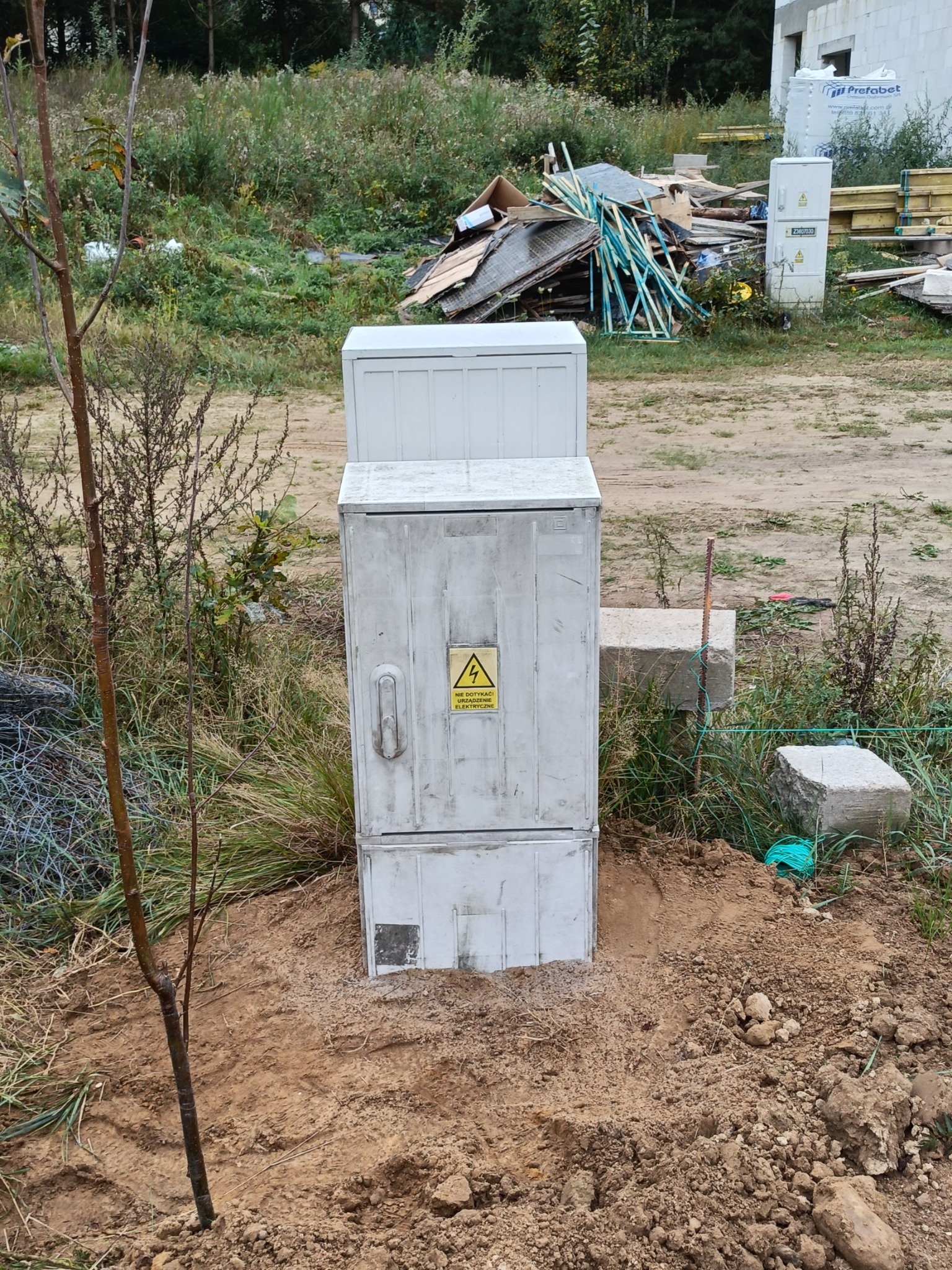 Szara skrzynka elektryczna z żółtą naklejką ostrzegawczą na tle placu budowy z widocznymi materiałami budowlanymi i młodym drzewkiem.