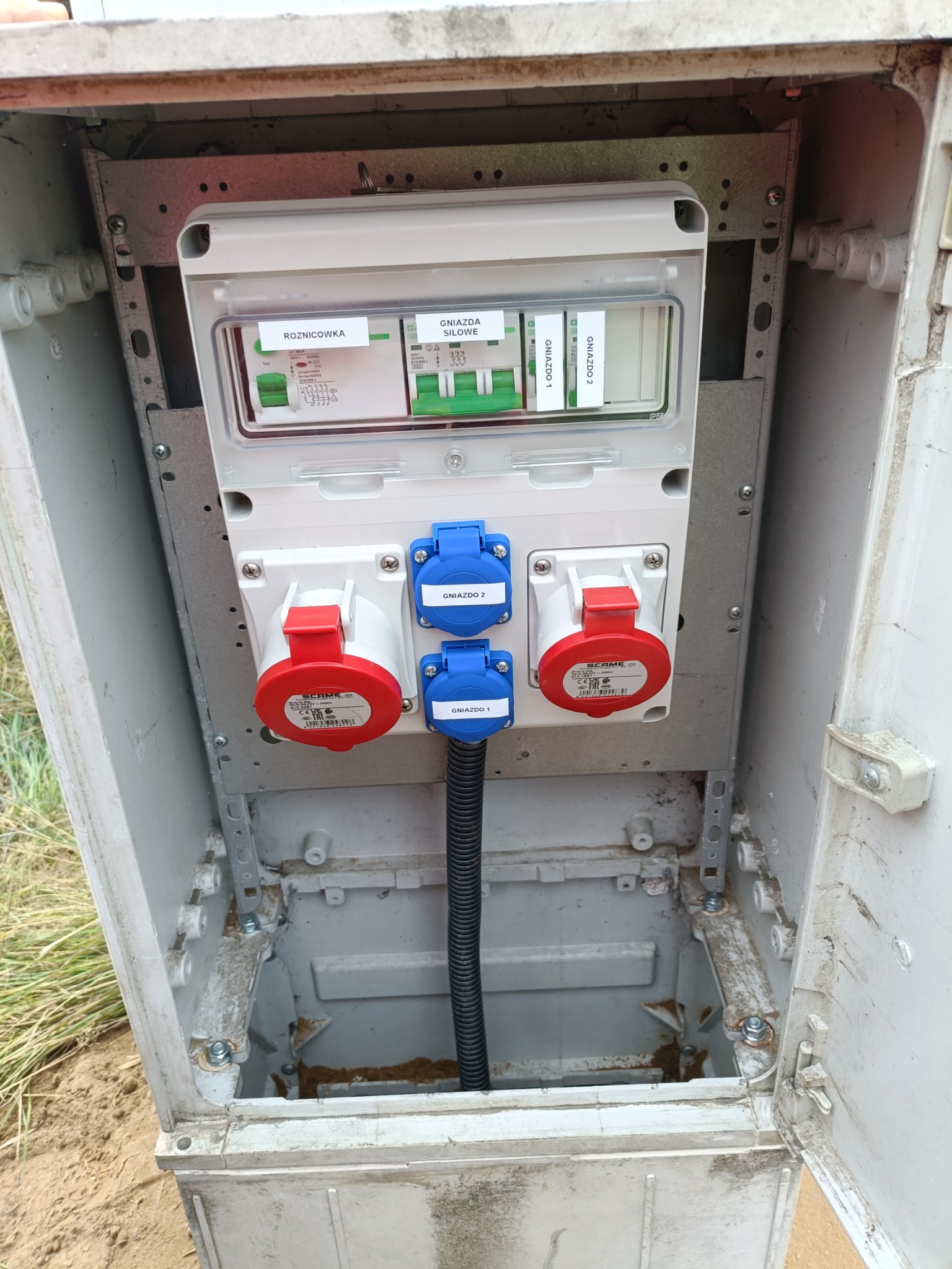 Otwarta skrzynka elektryczna z bezpiecznikami, wyłącznikami i gniazdami siłowymi w kolorach czerwonym i niebieskim, widoczne okablowanie i elementy montażowe.