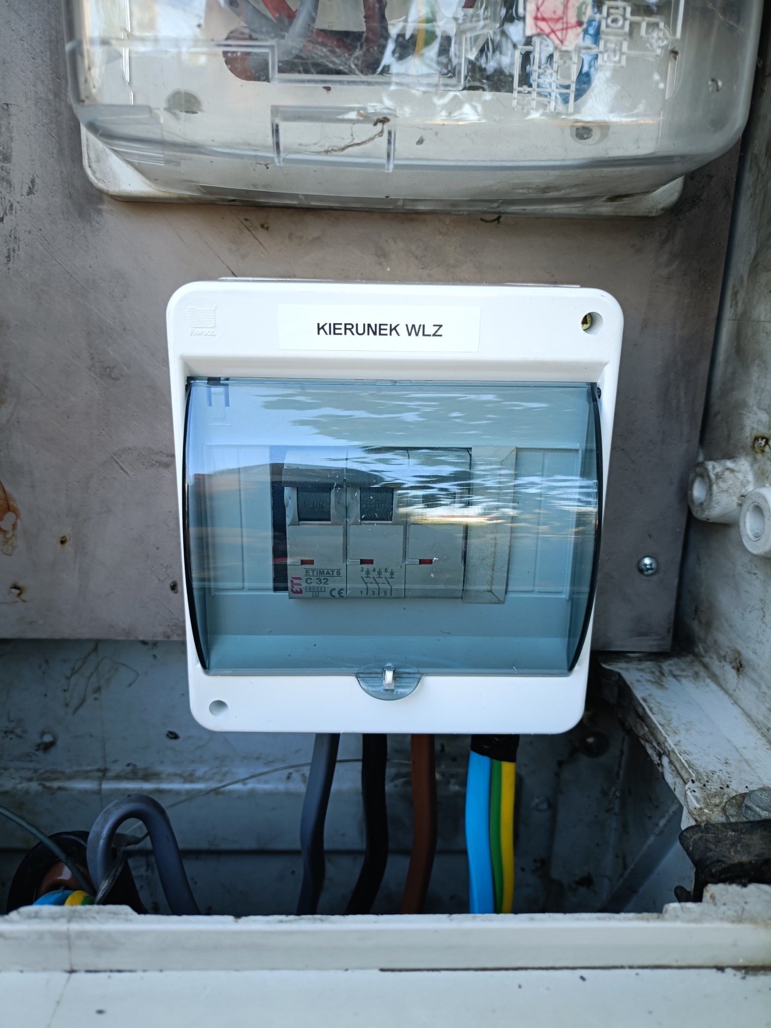 Skrzynka elektryczna z bezpiecznikami za przezroczystą osłoną, z naklejką 'KIERUNEK WLZ' i widocznymi przewodami.