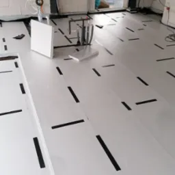 Białe płyty izolacyjne podłogowe układane w surowym wnętrzu, z czarnymi taśmami mocującymi, widoczne instalacje hydrauliczne i elektryczne, ściany z betonu komórkowego.