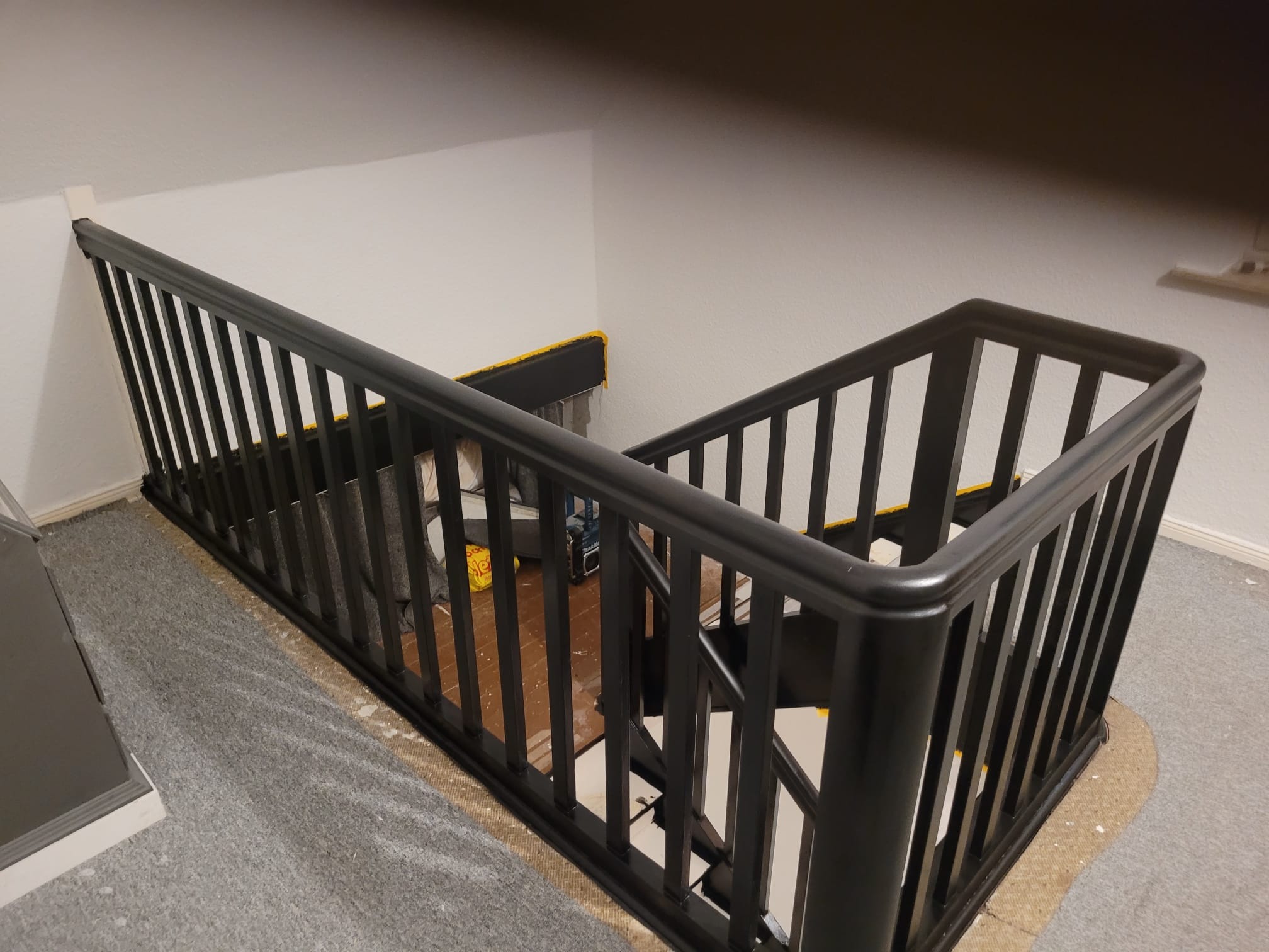 Czarna, metalowa balustrada schodowa z pionowymi szczebelkami, zamontowana na klatce schodowej w trakcie prac wykończeniowych, widoczne zabezpieczenia na schodach i podłodze.