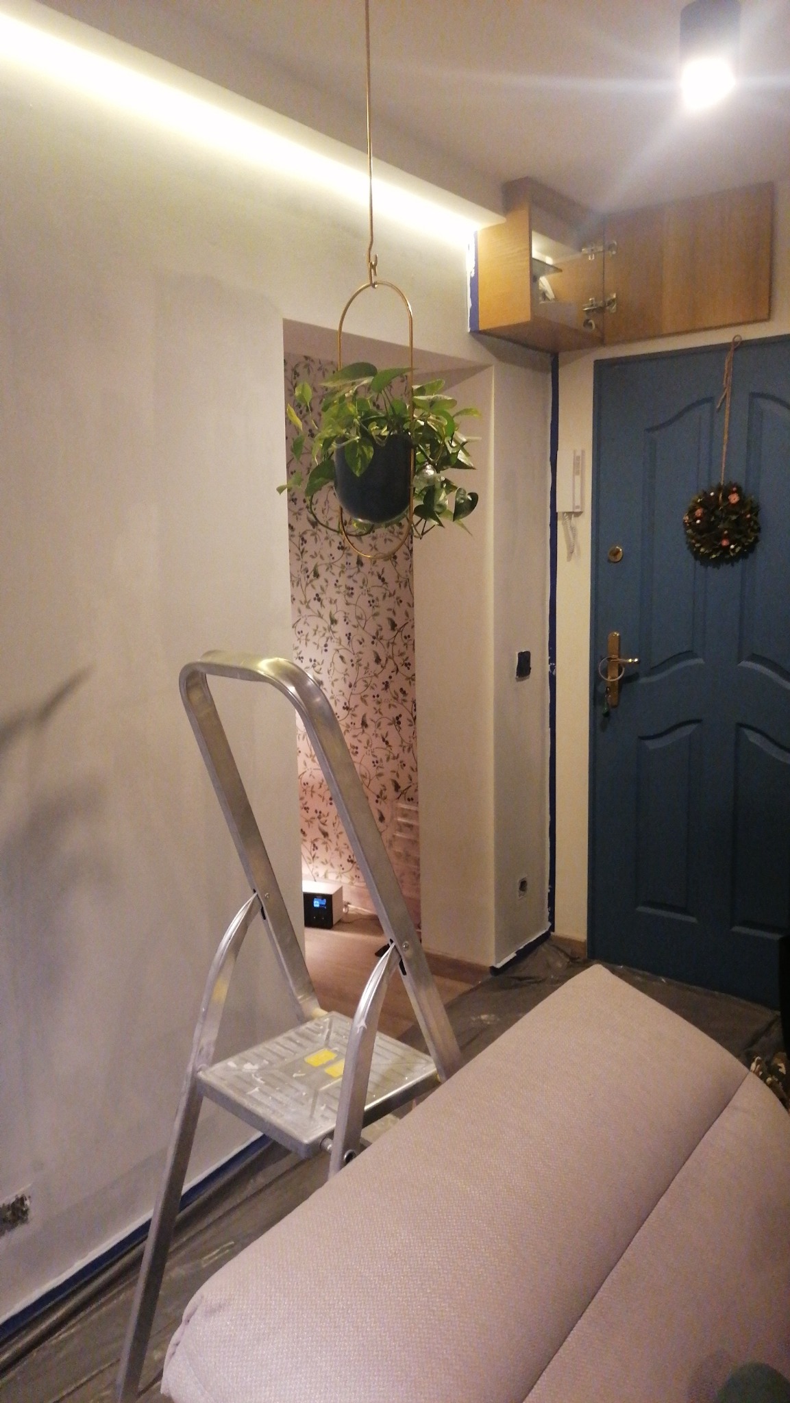 Ściana po szpachlowaniu, z widoczną drabiną, wiszącą rośliną w ozdobnym kwietniku i fragmentem mebla, przygotowana do dalszych prac wykończeniowych.