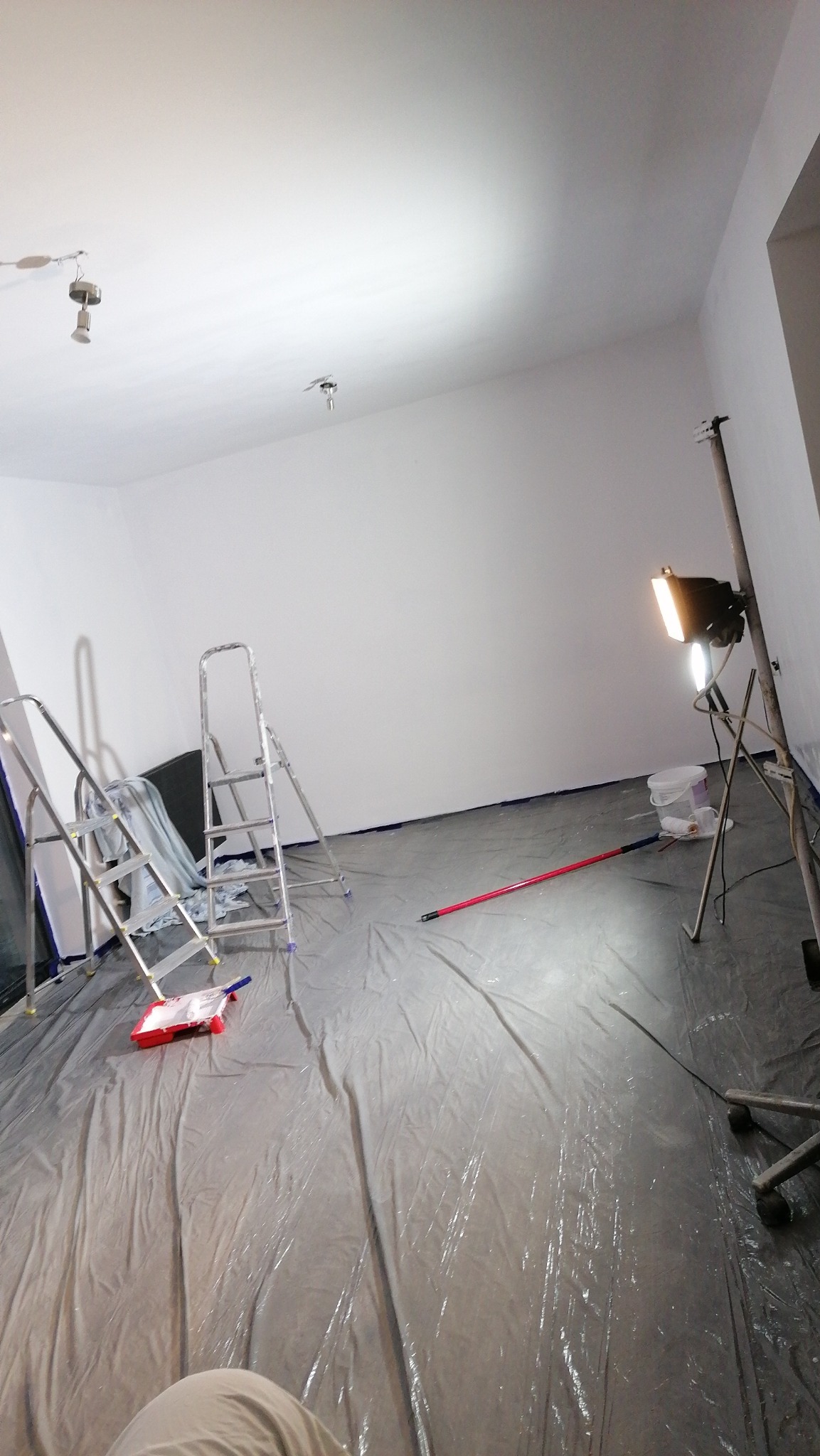 Przygotowane do malowania wnętrze z rozłożoną folią ochronną, dwoma drabinami, wałkiem malarskim w kuwecie, teleskopowym kijem do wałka, wiadrem z farbą i lampą halogenową na statywie.