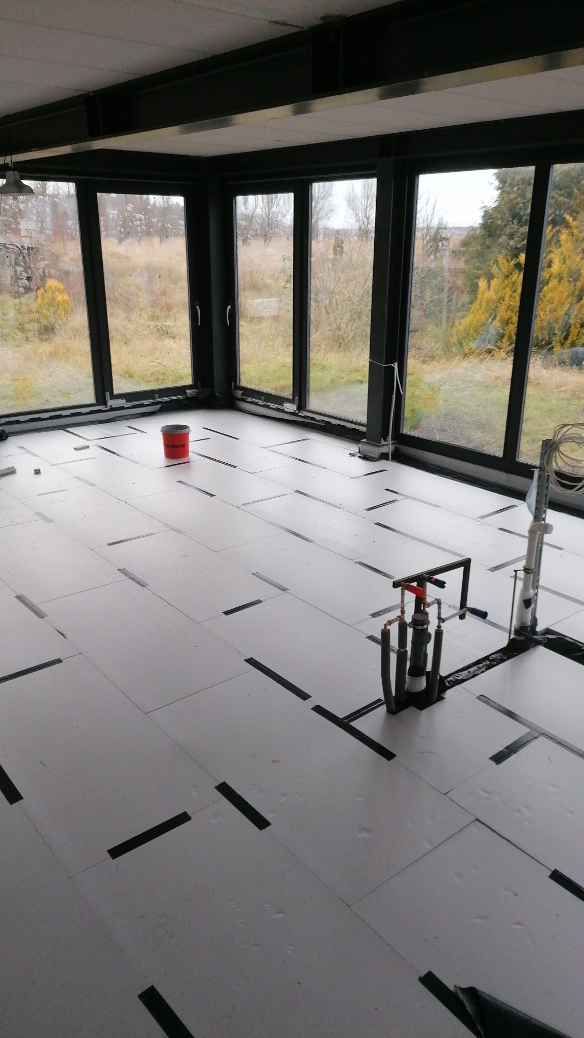 Przygotowanie podłoża podłogowego z płyt izolacyjnych, zabezpieczonych taśmą, w przeszklonym pomieszczeniu w Szczecinie, widok na zewnątrz przez okna.