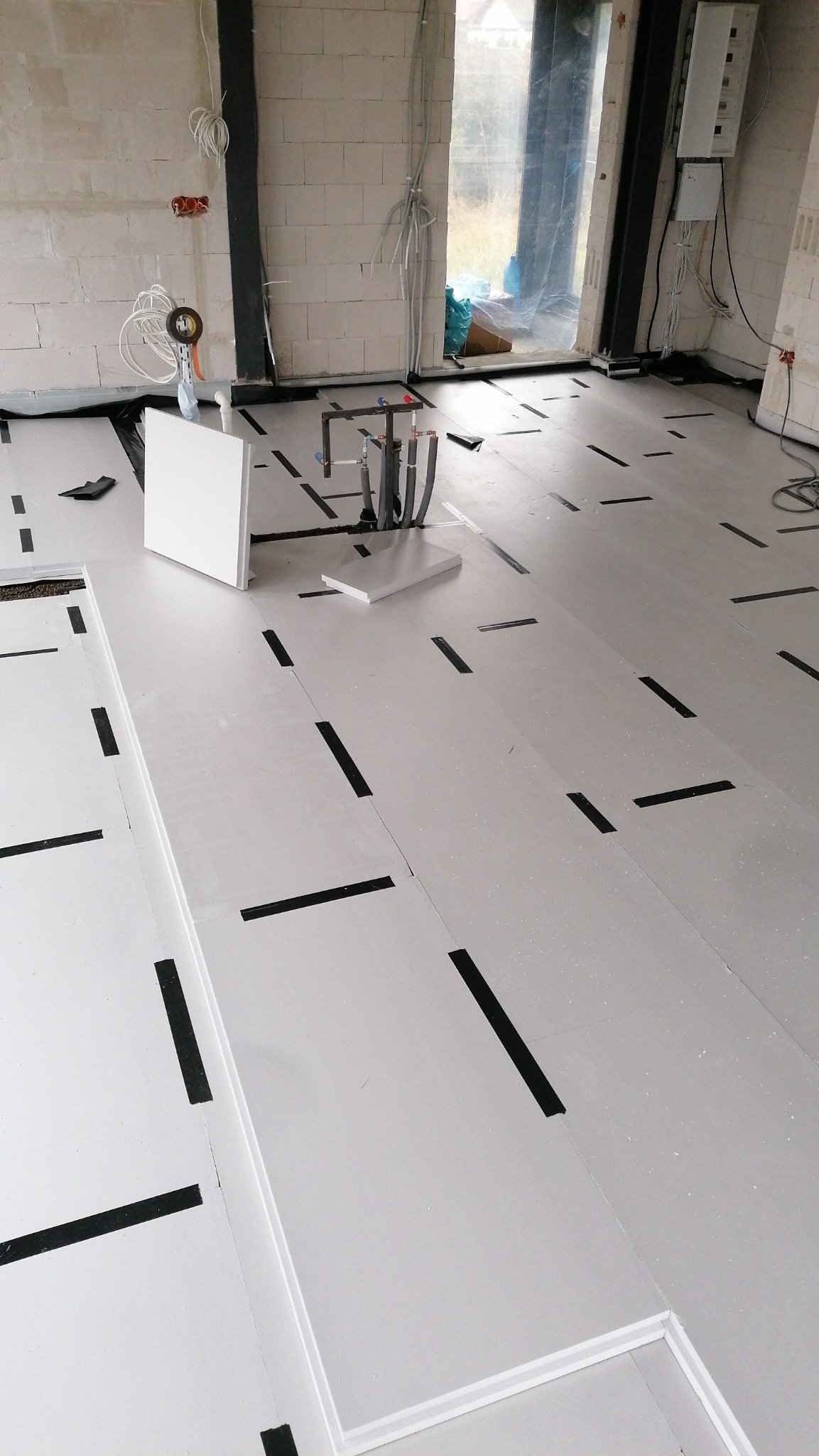 Białe płyty izolacyjne podłogowe układane w surowym wnętrzu, z czarnymi taśmami mocującymi, widoczne instalacje hydrauliczne i elektryczne, ściany z betonu komórkowego.