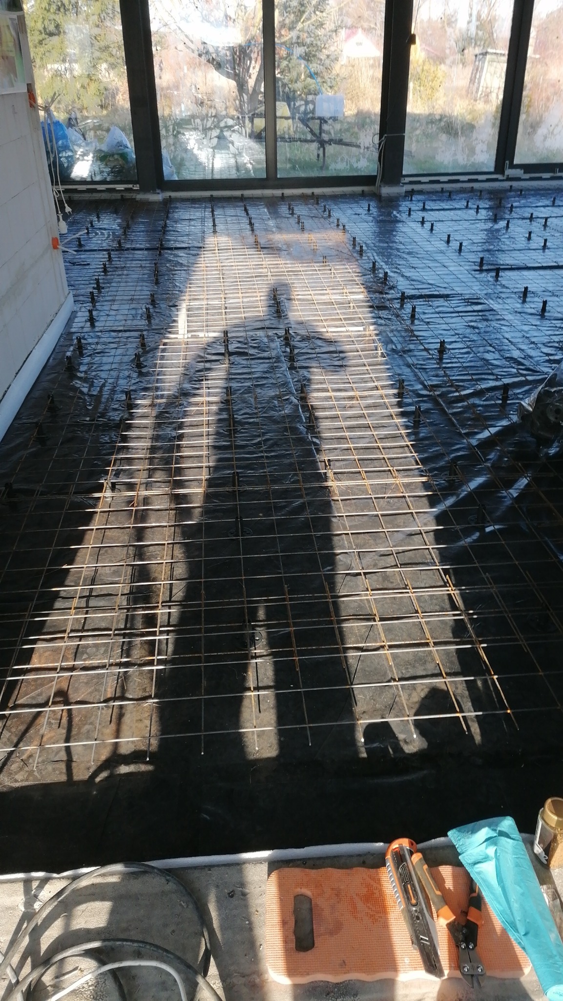 Przygotowanie podłoża pod wylewkę: siatka zbrojeniowa na folii izolacyjnej, widoczne narzędzia i cień osoby wykonującej prace w słoneczny dzień wewnątrz budynku.