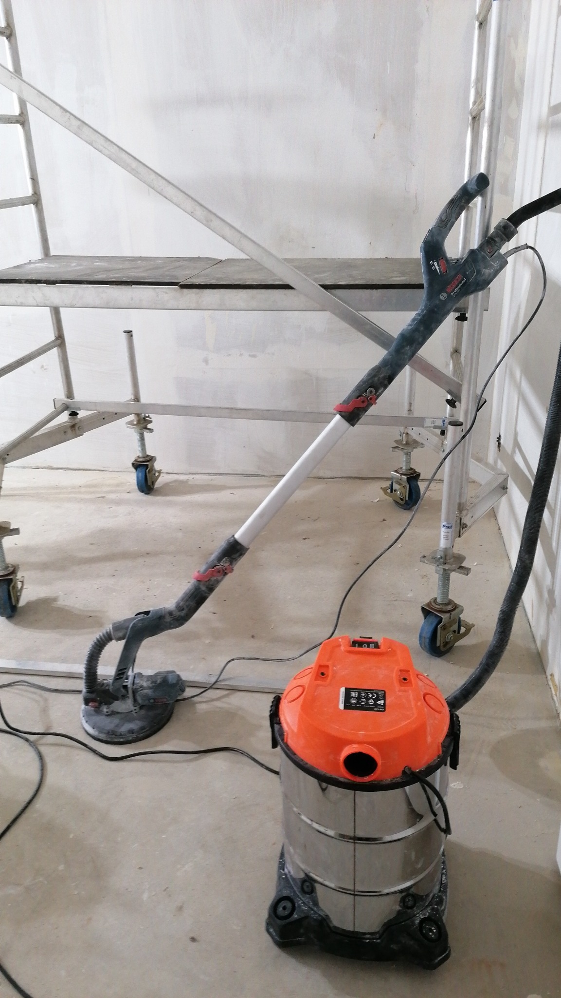 Szlifierka do gładzi gipsowych typu 'żyrafa' podłączona do odkurzacza przemysłowego na tle rusztowania w trakcie prac wykończeniowych.