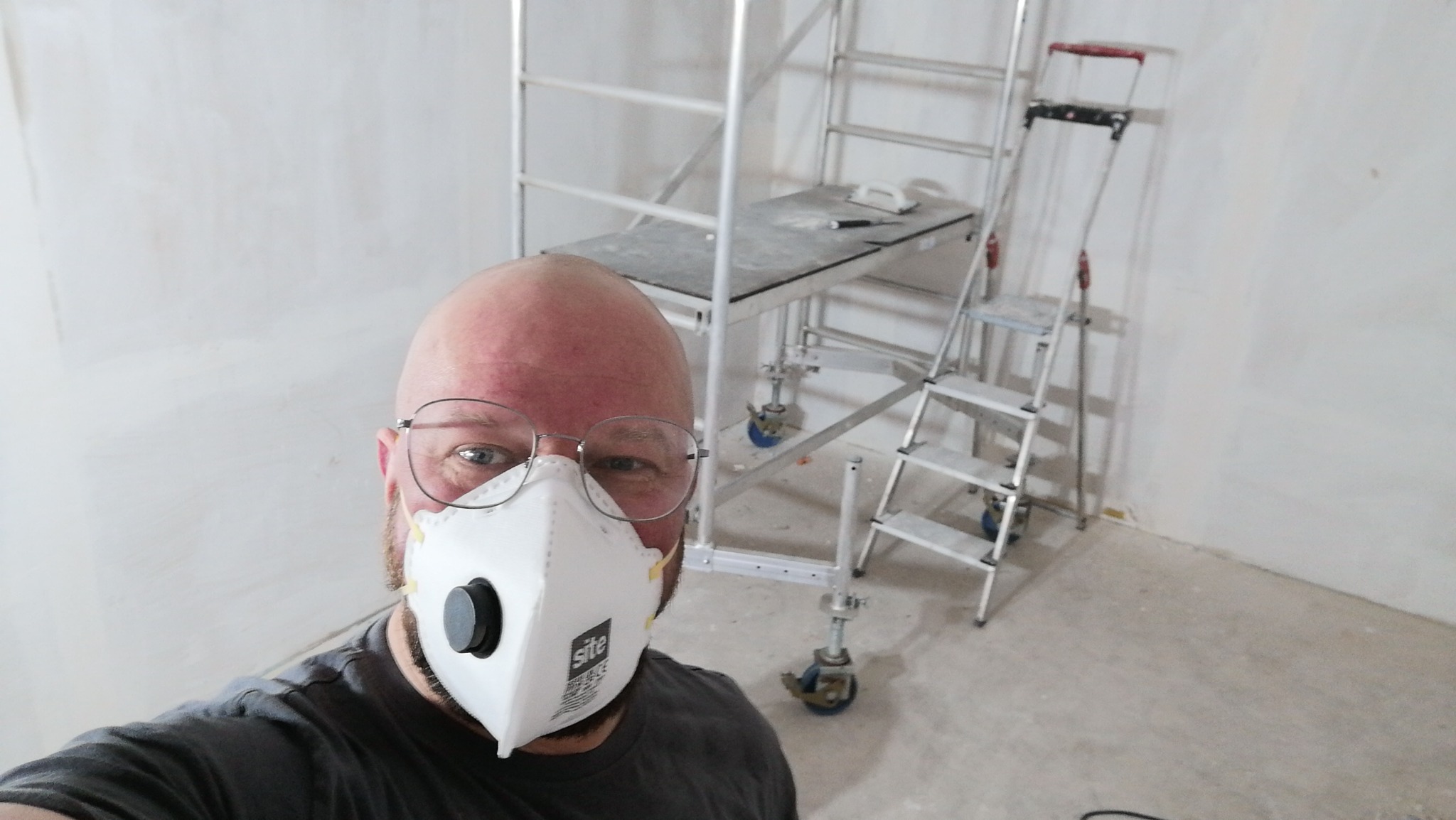 Autoportret mężczyzny w okularach i masce ochronnej na tle ściany po nałożeniu gładzi, z widocznym rusztowaniem i drabinką.