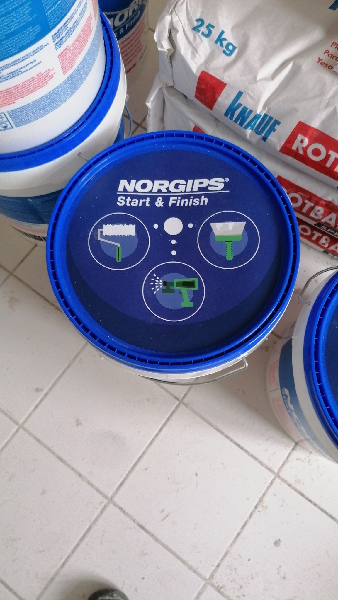 Wysoki kąt na wiadro z niebieską pokrywą Norgips Start & Finish z piktogramami wałka, szpachelki i pistoletu natryskowego, obok worka Knauf Rotband i innych wiader na tle płytek podłogowych.
