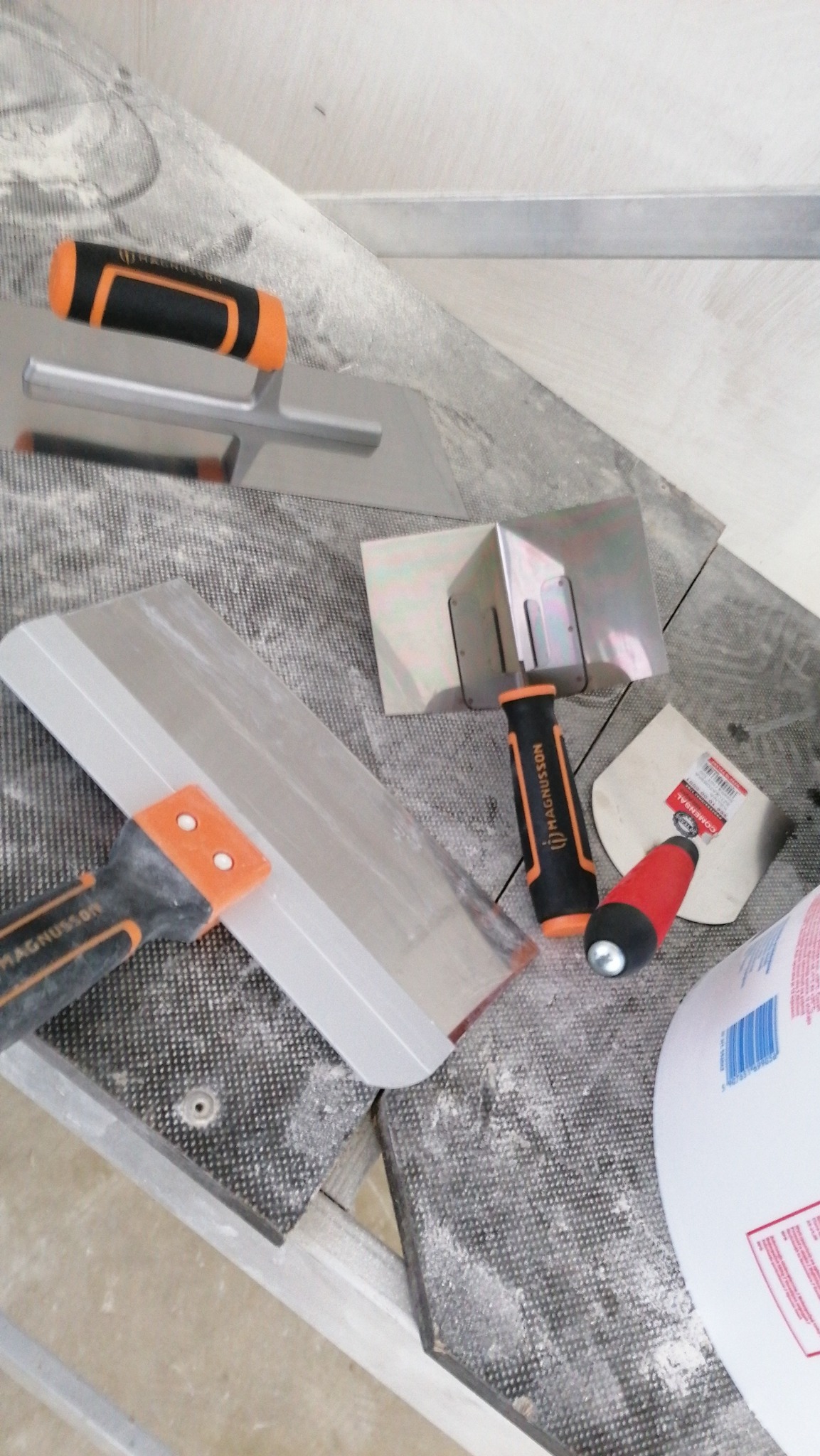 Zestaw narzędzi do nakładania gładzi: paca prosta Magnussona, paca do narożników, szpachelka i wiadro z masą szpachlową, leżące na powierzchni pokrytej pyłem.