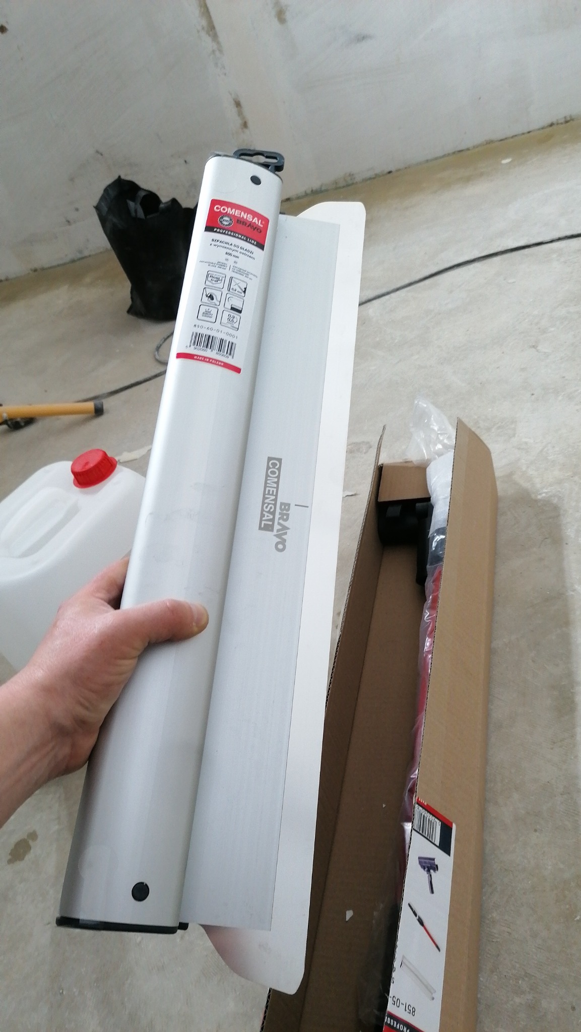 Ręka trzyma aluminiową szpachlę COMENSAL Bravo z etykietą, obok rozpakowane akcesoria w kartonowym pudełku, w tle surowy stan ścian i podłogi.