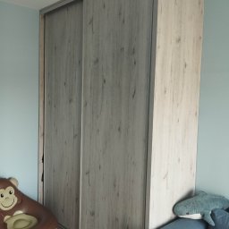 Artur Garbarczyk - Jasnoszara szafa przesuwna z drewnopodobnym wzorem, stojąca w pokoju dziecięcym, obok niebieskiej sofy z pluszowym rekinem i brązowego dmuchanego fotela w kształcie małpy.