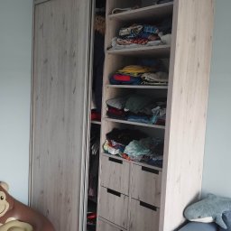 Artur Garbarczyk - Szafa wnękowa z przesuwnymi drzwiami i otwartymi półkami wypełnionymi ubraniami, widoczne dziecięce zabawki: dmuchany fotel małpa i pluszowy rekin.