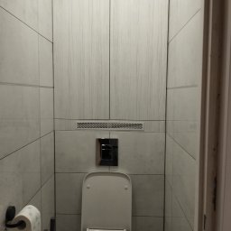 Artur Garbarczyk - Nowoczesna toaleta z ukrytą spłuczką i drewnopodobnymi płytkami na ścianach, widoczne akcesoria łazienkowe i środki czystości.