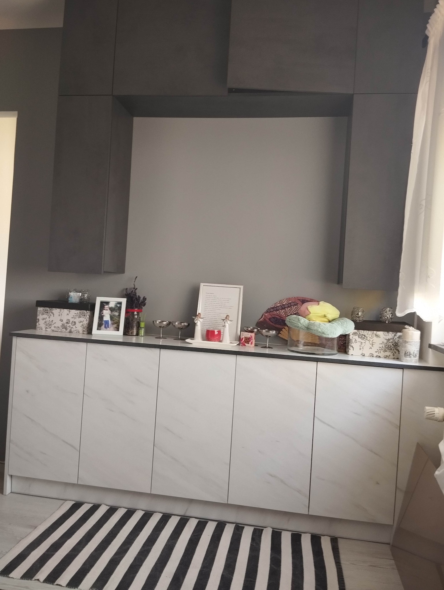Szara zabudowa meblowa z szafkami dolnymi z białymi frontami imitującymi marmur, udekorowana ramkami ze zdjęciami, świecznikami i innymi ozdobami, na podłodze czarno-biały dywanik.