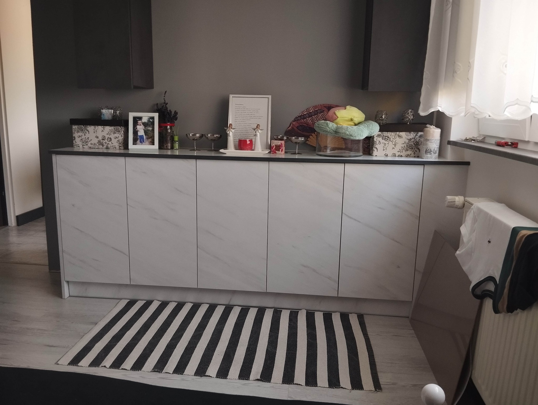 Biała komoda z marmurowym wzorem frontów, na której stoją ozdobne przedmioty, w tle szare ściany i ciemne szafki wiszące.