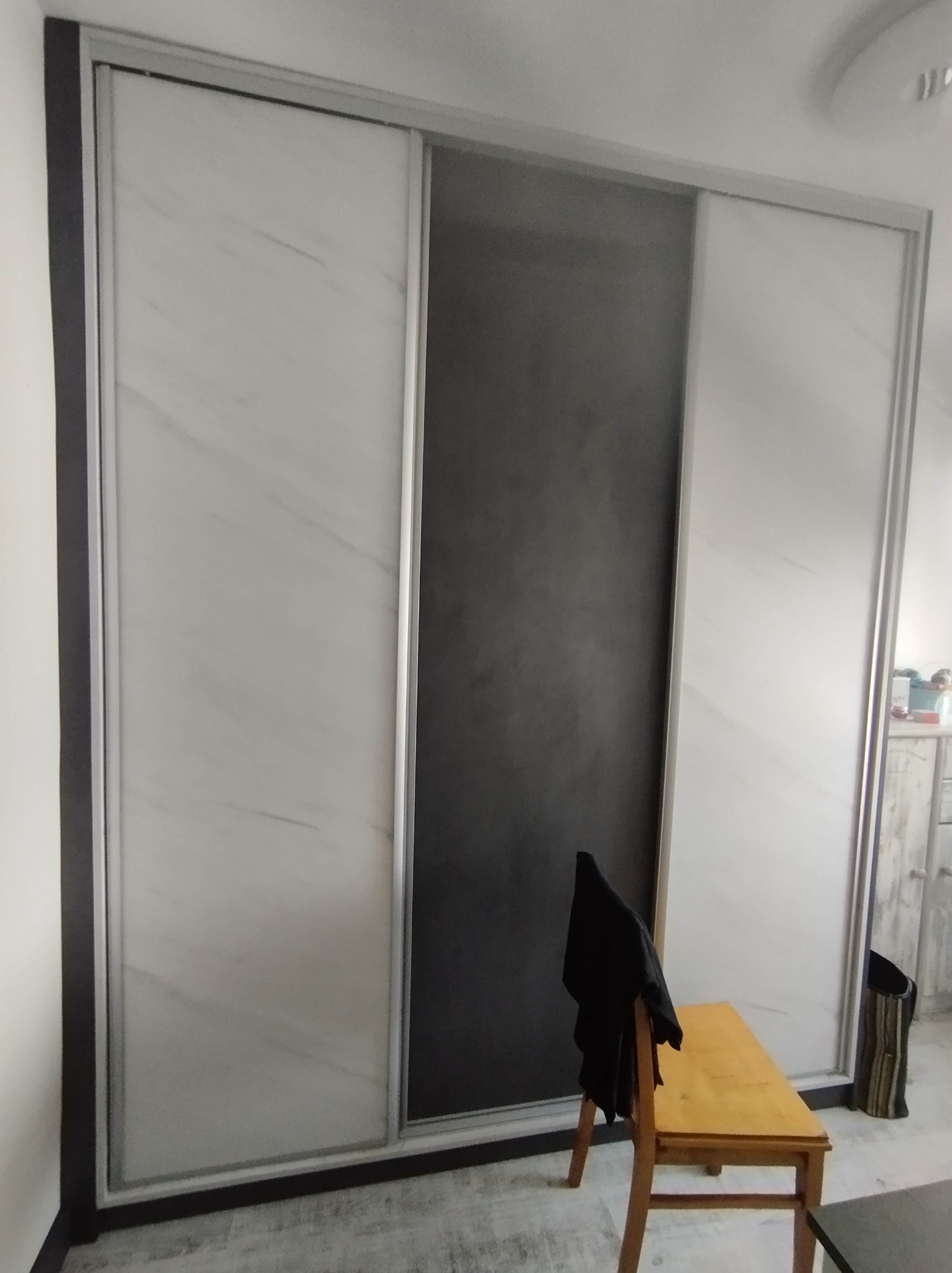 Trzydrzwiowa szafa wnękowa z przesuwnymi drzwiami: białe panele imitujące marmur po bokach i grafitowy panel pośrodku, widoczny drewniany stołek z czarną kurtką.