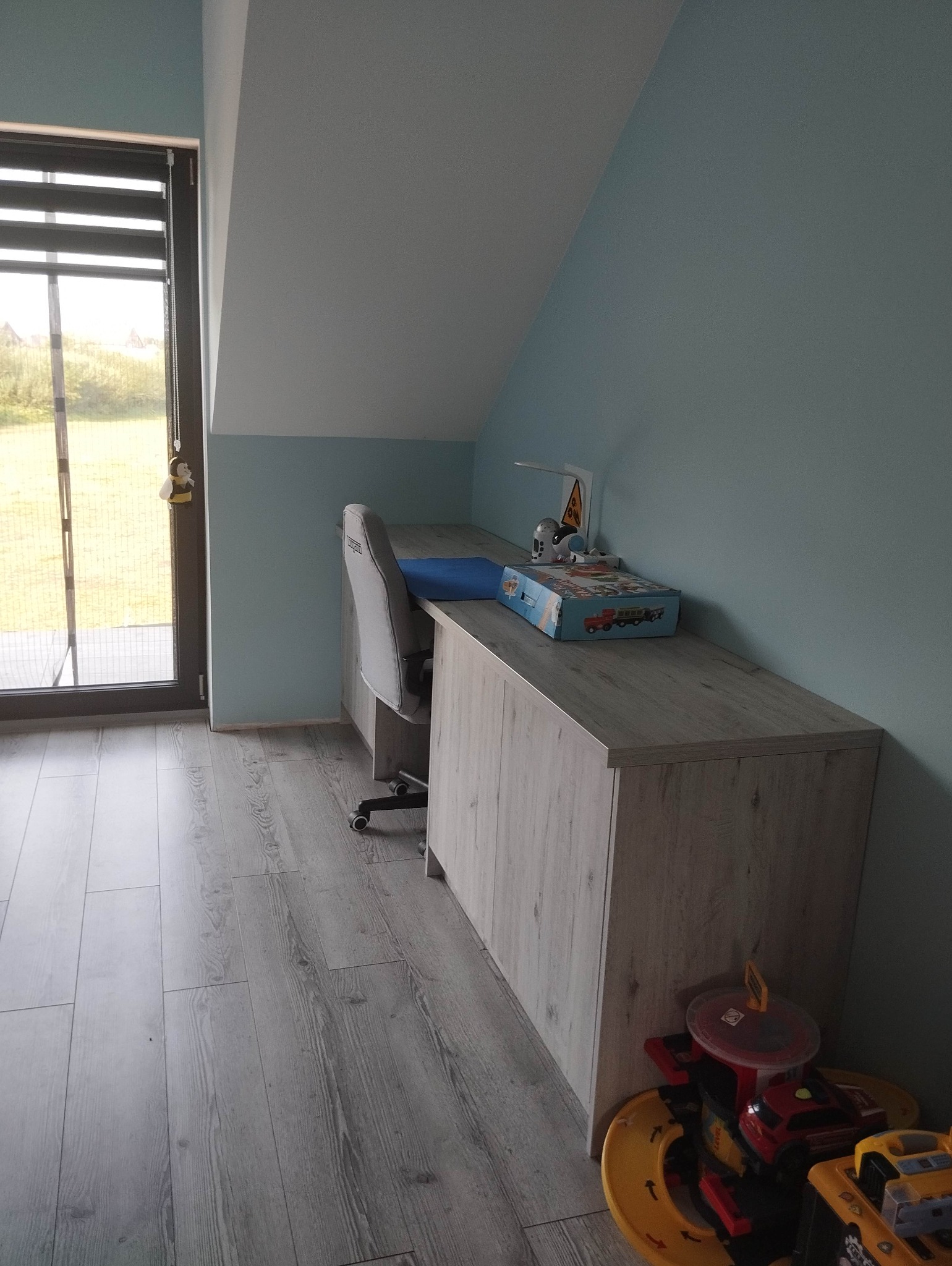 Pokój dziecięcy na poddaszu z jasną podłogą, biurkiem z szufladami w kolorze jasnego drewna, krzesłem biurowym i zabawkami pod biurkiem.