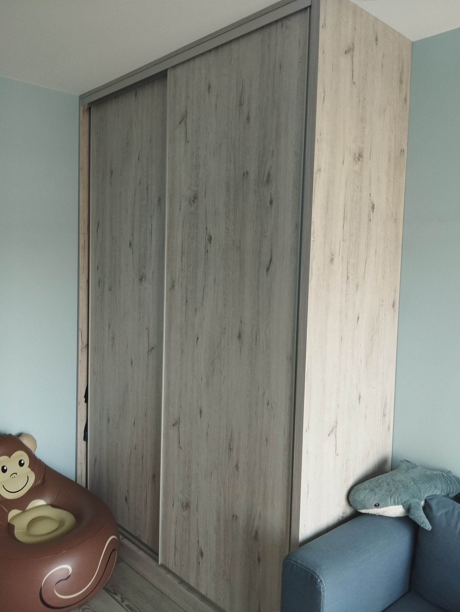 Jasnoszara szafa przesuwna z drewnopodobnym wzorem, stojąca w pokoju dziecięcym, obok niebieskiej sofy z pluszowym rekinem i brązowego dmuchanego fotela w kształcie małpy.