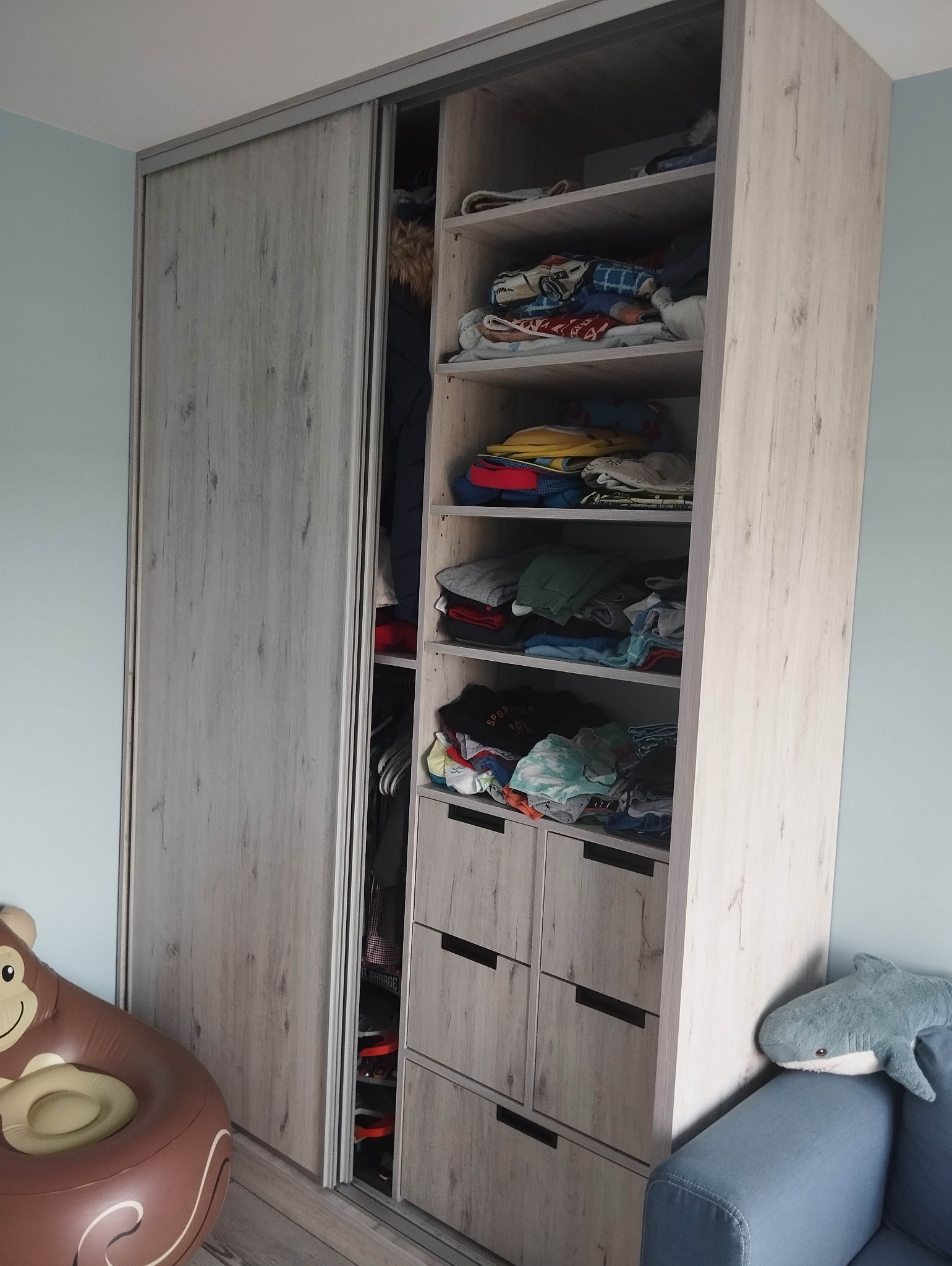 Szafa wnękowa z przesuwnymi drzwiami i otwartymi półkami wypełnionymi ubraniami, widoczne dziecięce zabawki: dmuchany fotel małpa i pluszowy rekin.