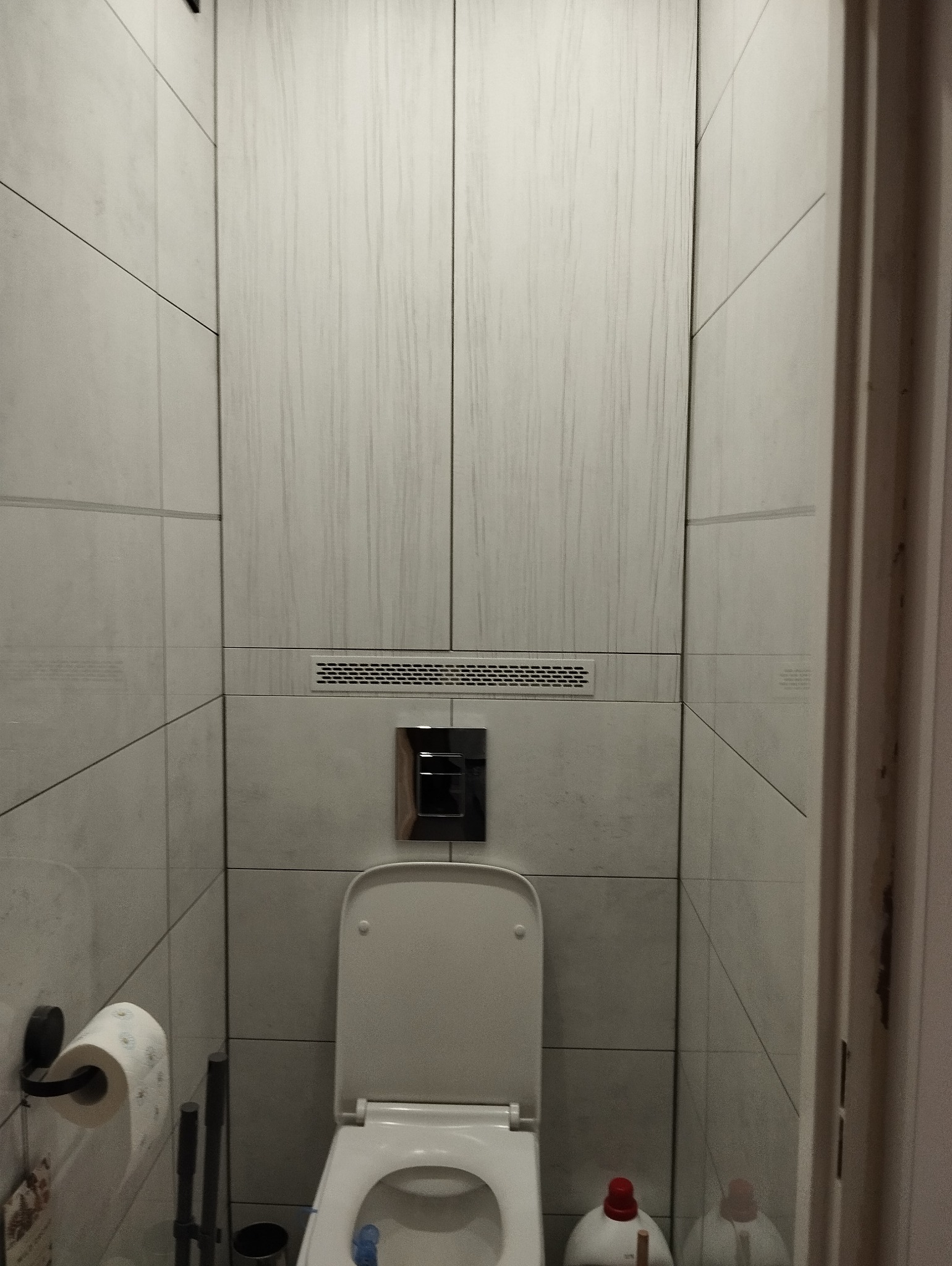 Nowoczesna toaleta z ukrytą spłuczką i drewnopodobnymi płytkami na ścianach, widoczne akcesoria łazienkowe i środki czystości.