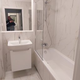 Hennadii Lytvyn - Wykończona łazienka z białymi płytkami imitującymi marmur, białą umywalką z szafką i wanną z parawanem, widoczne odbicie fotografa w lustrze.