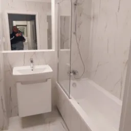 Wykończona łazienka z białymi płytkami imitującymi marmur, białą umywalką z szafką i wanną z parawanem, widoczne odbicie fotografa w lustrze.