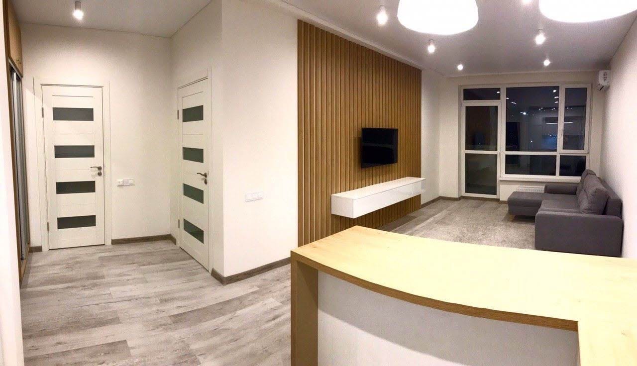 Jasne wnętrze mieszkania po remoncie: panele podłogowe imitujące drewno, ściana z drewnianych lameli za telewizorem, biała szafka RTV, szara sofa narożna pod oknem, dwie białe drzwi z czarnymi...