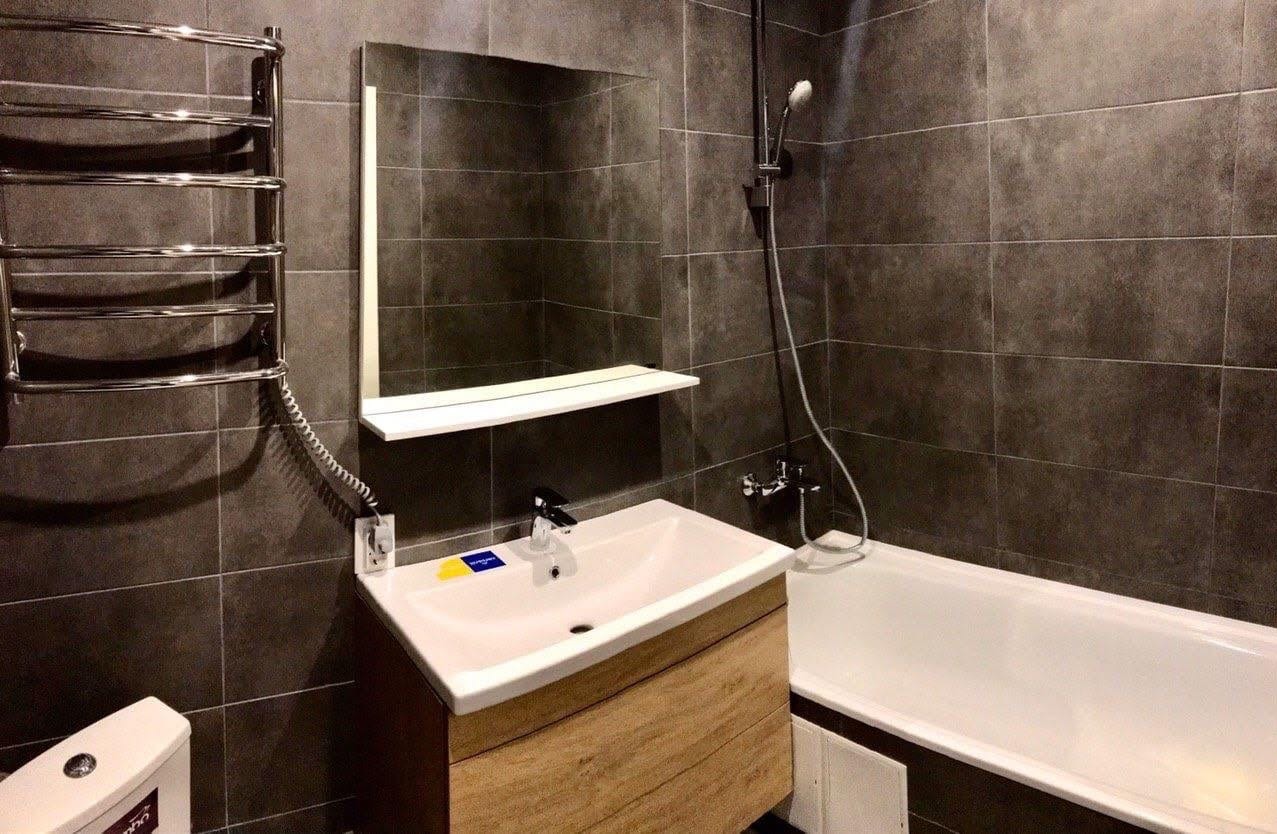 Nowoczesna łazienka z szarymi płytkami, wiszącą szafką umywalkową z drewnianym frontem, grzejnikiem drabinkowym i wanną z prysznicem.