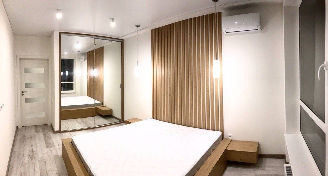 Nowoczesna sypialnia z drewnianą ścianą dekoracyjną, łóżkiem na niskiej platformie, szafą z lustrzanymi drzwiami przesuwnymi i klimatyzacją.